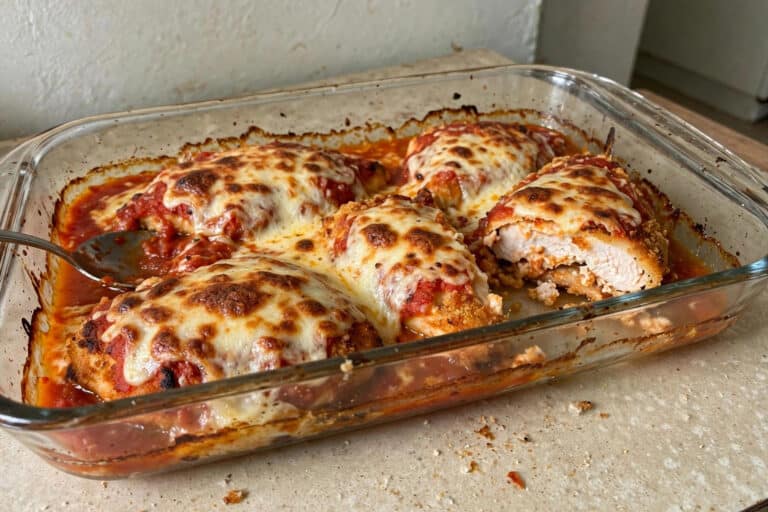 Refratário de vidro com filé de frango à parmegiana gratinado com mussarela e molho de tomate sobre a bancada da cozinha, um filé cortado mostrando o frango suculento